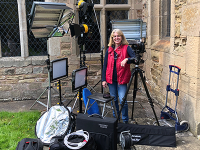 Woman stands with camera and lighting outside an old church building wearing pink top