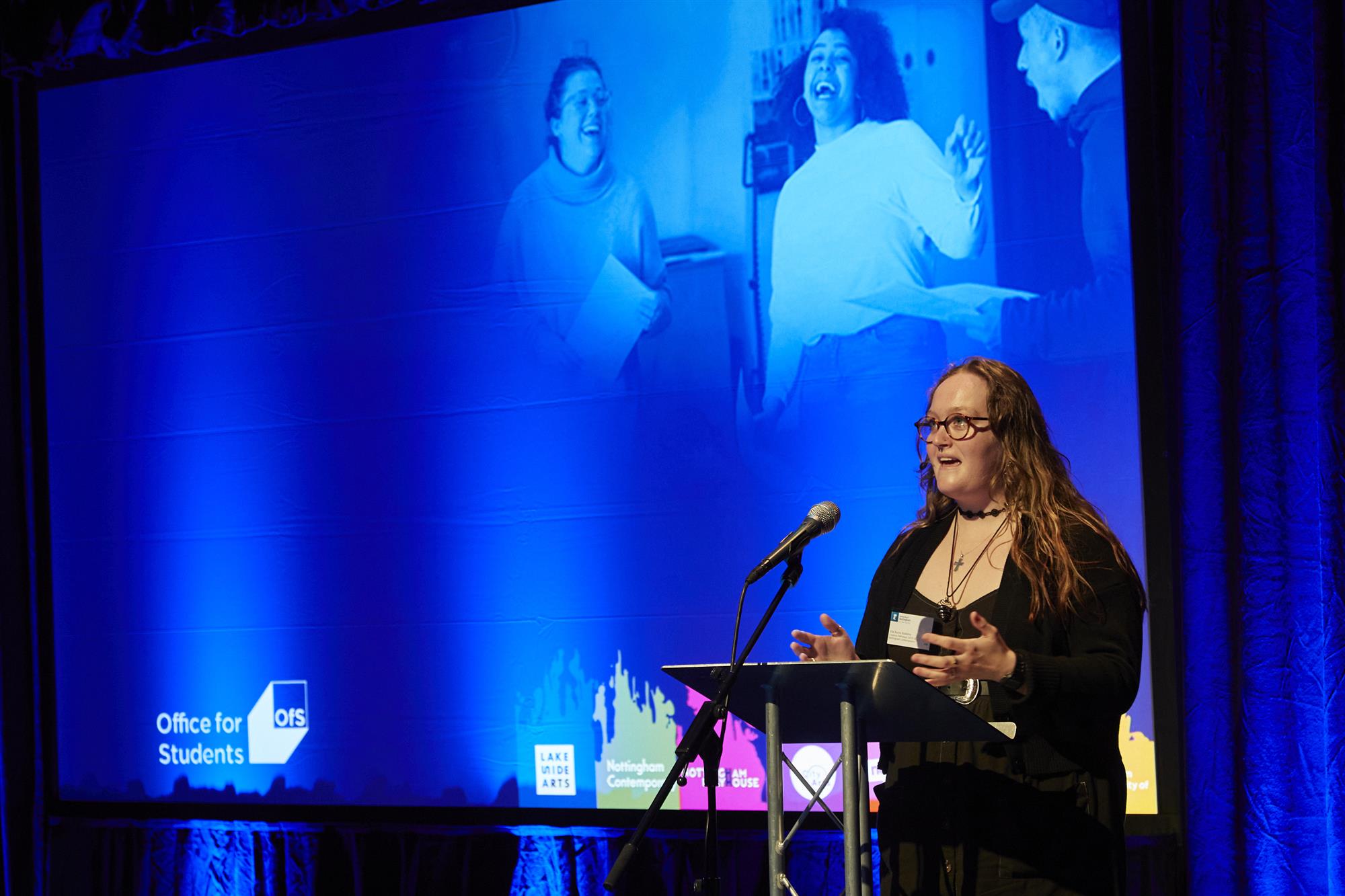 Poet Ella Burns Robbins stands on stage performing, with the blue Creative Pathways slides in the background