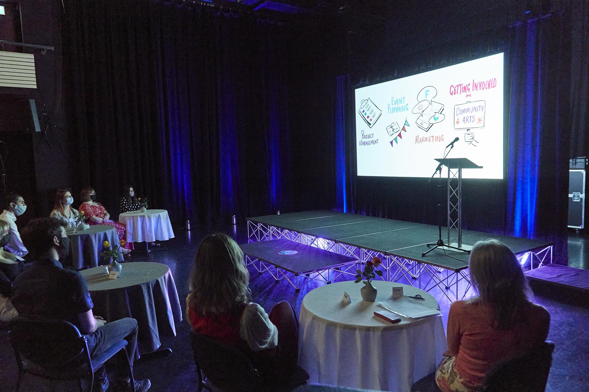 The seven interns sit at socially distanced tables, watching the animated video playing on stage