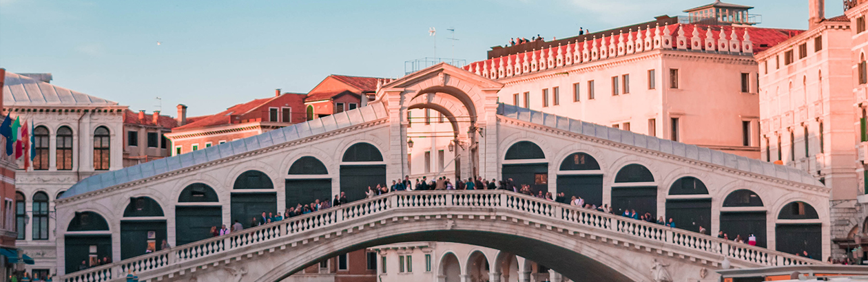 Photograph of tall buildings, a bridge across water with people standing on it and blue sky in Venice