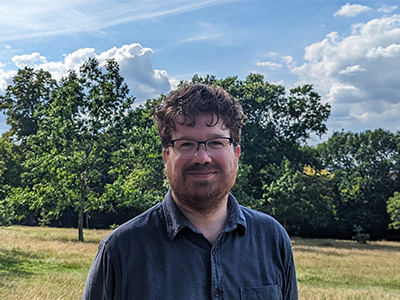 A man with brown hair and a brown beard and glasses stands in a green grassy space