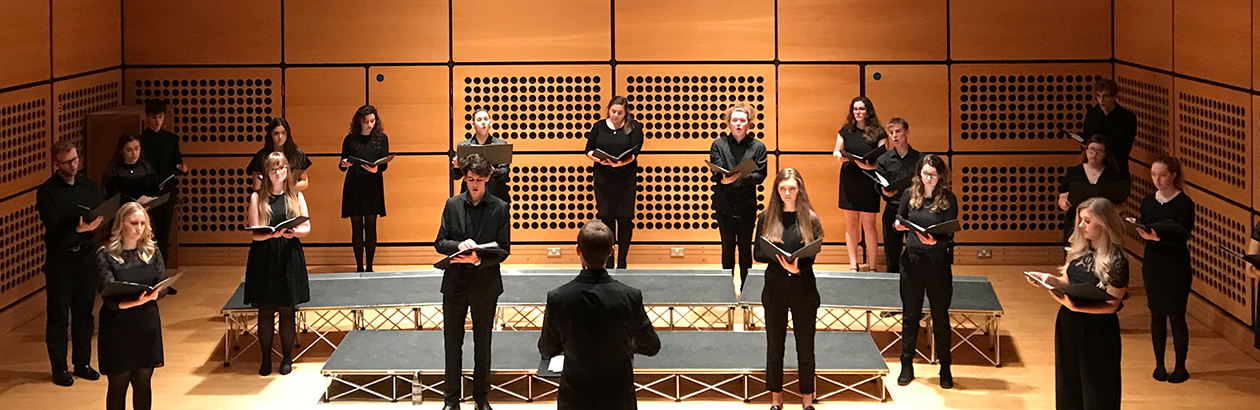 University of Nottingham Chamber Choir standing distances in the Djanogly Recital Hall. About 30 people stand wearing black against a brown wall