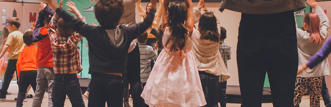 Photograph of a group of children facing a grown up and following instructions. All raising arms