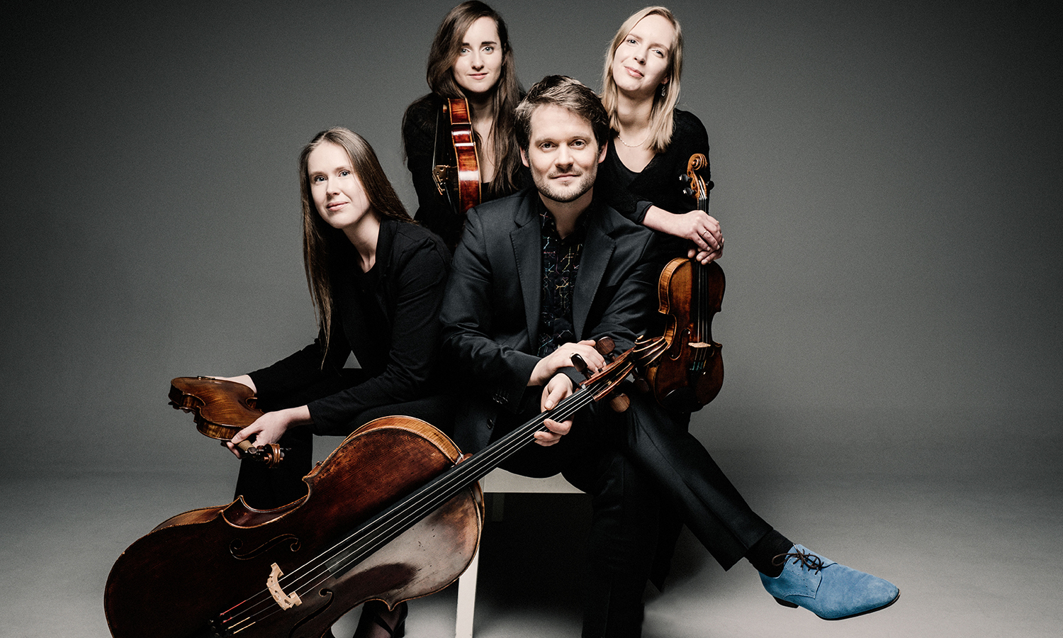 three females stand holding string instruments with a male musician in foreground with cello at base of feet