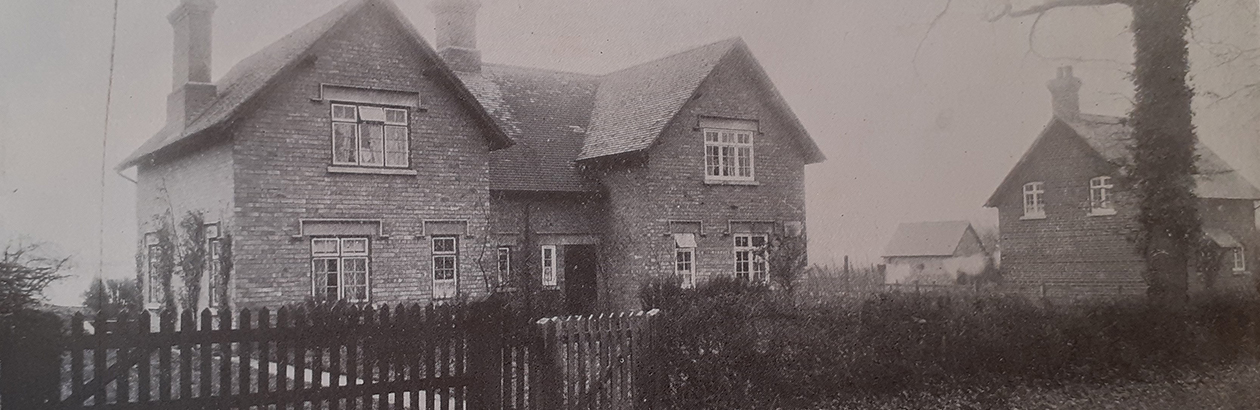 Photograph of the front of a house with a white fence and trees in front garden. Photo in black and white