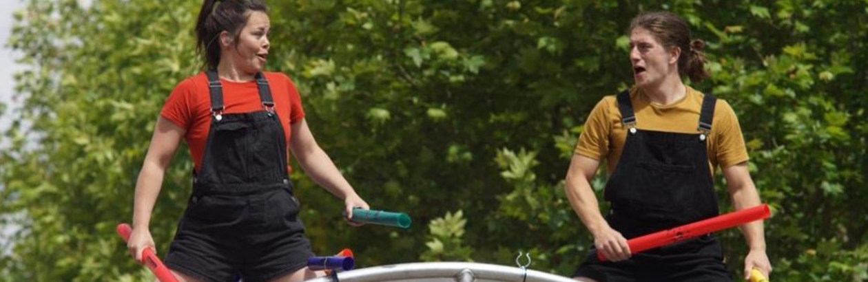 Two dancers looking at each other. Female on left dressed in red tshirt and black dungarees. Male dancer on right dressed in yellow tshirt and black dungarees. Holding instruments