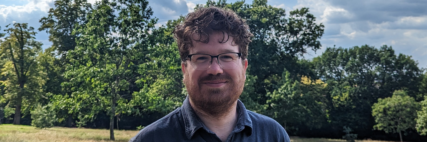 A man with brown hair and a brown beard stoood in a grassy area