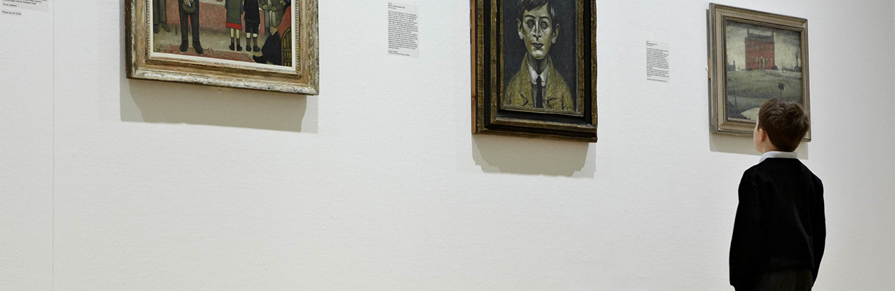 Photograph of a boy in gallery. Boy wears school uniform and observes Lowry paintings. He is looking at painting of a face.