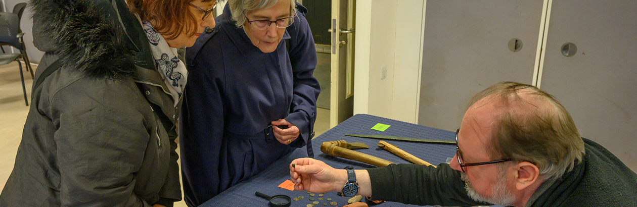 Photograph of two women observing an item on table with a man holding hands out holding item