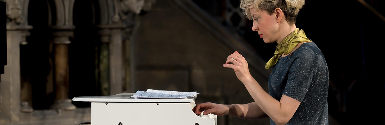 Photo of Xenia Pestova Bennet playing a miniature white piano wearing a grey top and green scarf